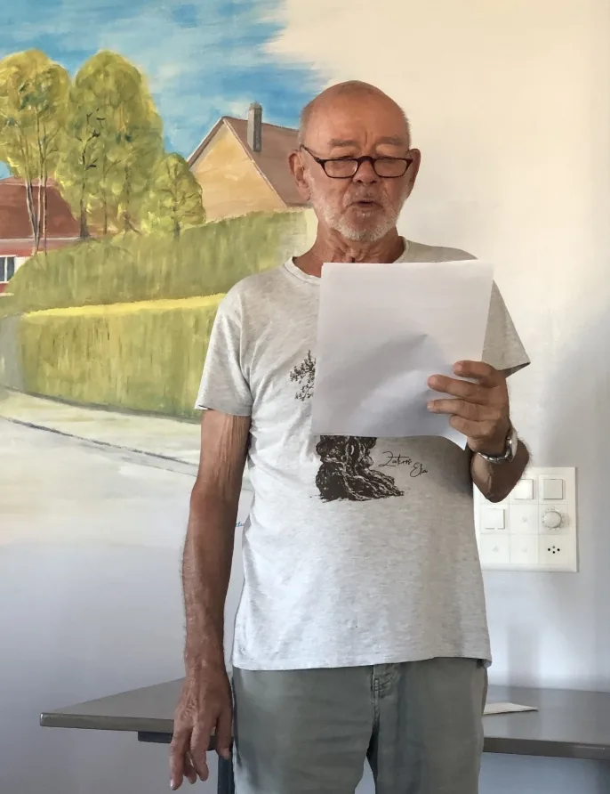 Claude Maier lisant un texte, debout devant une peinture murale