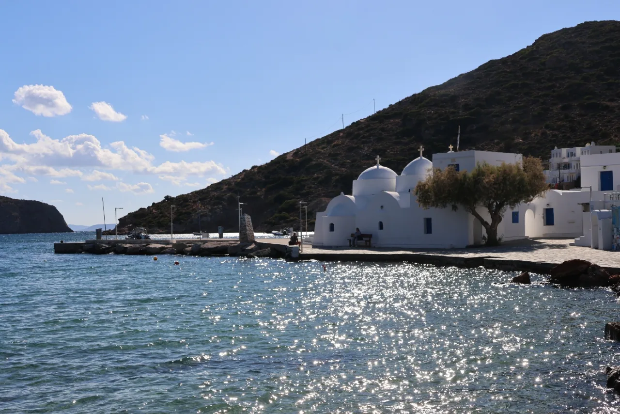 Baie de Sifnos, dans les Cyclades : chapelle blanche aux coupoles au bord d'une mer scintillante, sous une colline aride